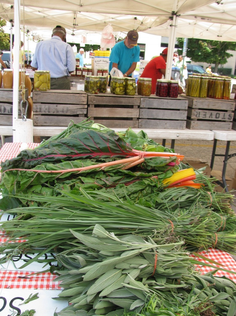 Štand s obiljem zelenja: blitva u svim bojama (tzv. rainbow chard), ružmarin, kadulja (i onda redom dalje, bez slike: origano, bosiljak, menta, peršin, rikola, kelj, i drugo uobičajeno svježe bilje) . U pozadini, ukiseljeno povrće.